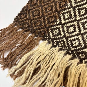 May include: A woven textile featuring a geometric diamond pattern in shades of dark brown, medium brown, and cream. The fabric edge is finished with long, thick fringe tassels in alternating colors of light brown and cream throughout the bottom.