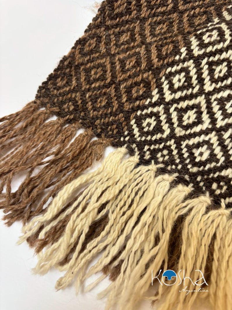 May include: A woven textile featuring a geometric diamond pattern in shades of dark brown, medium brown, and cream. The fabric edge is finished with long, thick fringe tassels in alternating colors of light brown and cream throughout the bottom.