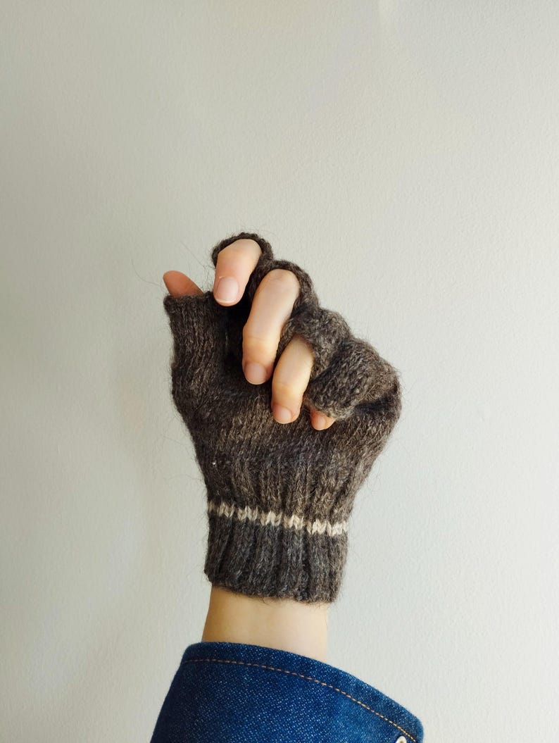 Puede incluir: Un guante sin dedos de color marr&oacute;n oscuro con un pu&ntilde;o acanalado y una fina raya horizontal de color crema. La textura del tejido es visible en toda la prenda, que cubre la palma y el dorso de la mano dejando los dedos al descubierto.