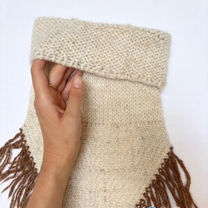 May include: A cream-coloured knit neck warmer featuring a folded top edge and a woven lower section with long brown fringe trim. The bottom right corner displays the text Kosha Argentina in a white script font with a blue circular logo.