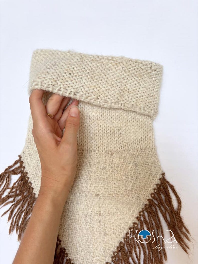 May include: A cream-coloured knit neck warmer featuring a folded top edge and a woven lower section with long brown fringe trim. The bottom right corner displays the text Kosha Argentina in a white script font with a blue circular logo.