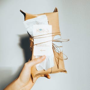 以下が含まれることがあります： 茶色のクラフト紙で包まれ、細い麻紐と水色のワックスシールで留められた長方形の小包。前面には白い長方形のタグが付けられており、オレンジ色でKoshaという文字と、その下に小さな印刷されたテキストが記載されています。