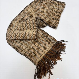 May include: A woven scarf featuring a repeating diamond pattern in shades of beige, brown, and dark gray. The scarf has a textured appearance and is finished with long, loose fringe at one end. The logo Kosha Argentina is visible.
