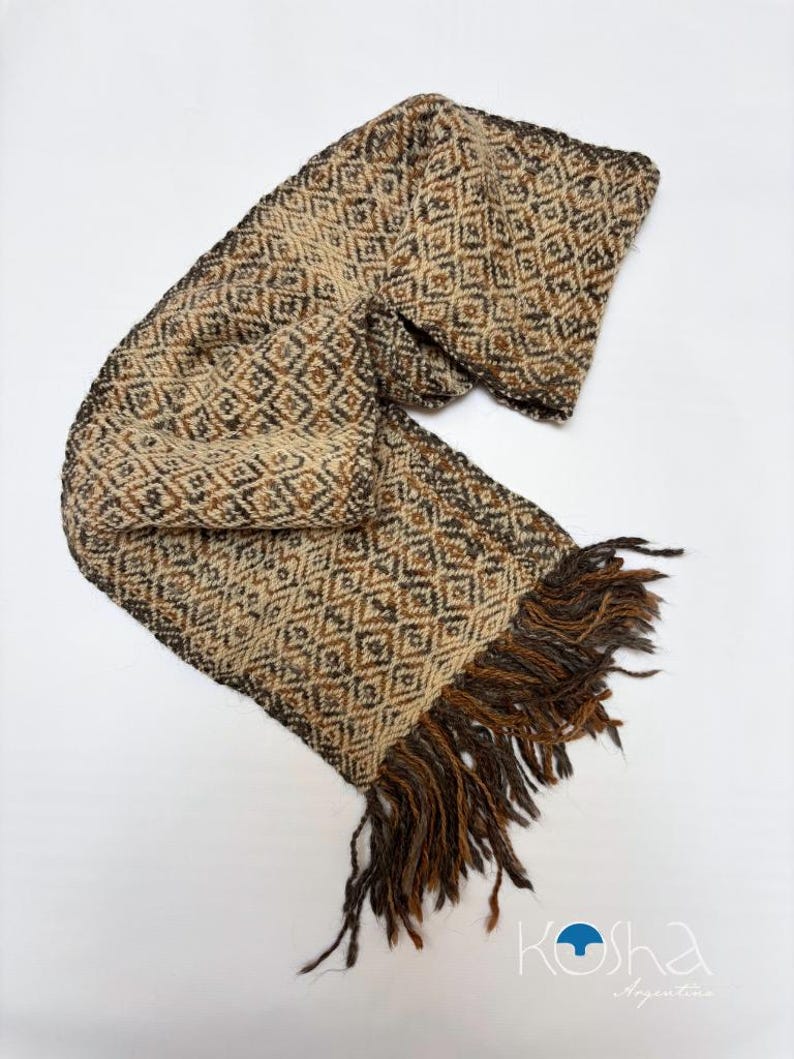 May include: A woven scarf featuring a repeating diamond pattern in shades of beige, brown, and dark gray. The scarf has a textured appearance and is finished with long, loose fringe at one end. The logo Kosha Argentina is visible.