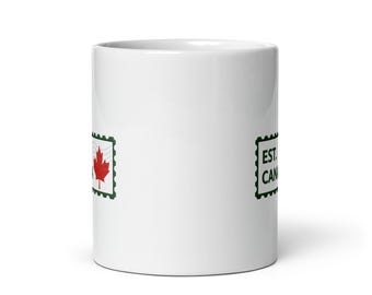 May include: White ceramic mug with a red maple leaf stamp design and the text "EST. CAN". The mug has a glossy finish and a standard handle. The design is printed on both sides of the mug.
