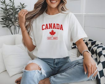 Pode incluir: Camiseta branca com a palavra "CANADA" em vermelho, uma folha de bordo vermelha e "EST. 1867". A camiseta é combinada com jeans azuis. O design celebra o Canadá.