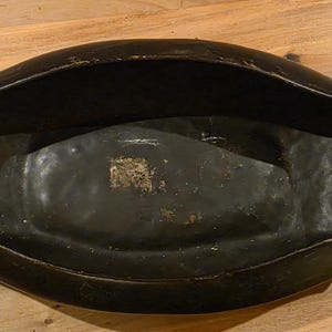 May include: A dark, oblong wooden bowl with a curved design. The bowl has a black finish with visible wear and tear, suggesting age. The interior has a slightly lighter, textured surface.
