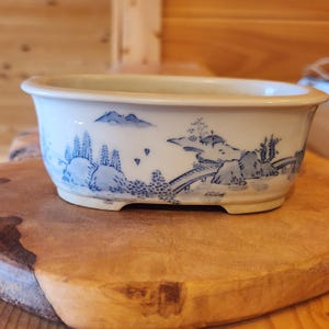 May include: A white ceramic bonsai pot with a blue landscape design. The oval-shaped pot features a detailed illustration of trees, mountains, and a bridge. The pot has a slightly raised base and a smooth, glossy finish.