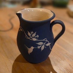 May include: A navy blue ceramic pitcher with a white interior and handle. The pitcher features a white hummingbird and floral design. The pitcher is likely used for serving beverages and adds a decorative touch to any table setting.