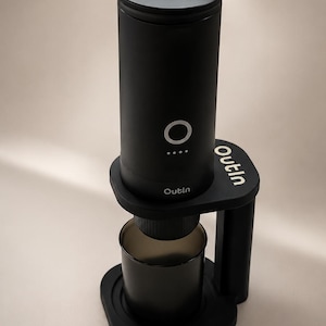 May include: A matte black portable espresso maker with a cylindrical body, a circular power button, and four indicator lights. The device features the brand name Outin printed on the base and the main unit, and it sits on a matching black stand.