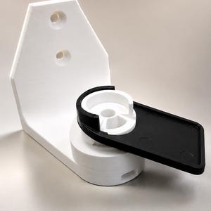 May include: A white wall-mounted bracket featuring a vertical backplate with two circular mounting holes. A cylindrical base supports a rotating mechanism with a black rectangular arm and a circular white cup-like holder attached to the front.