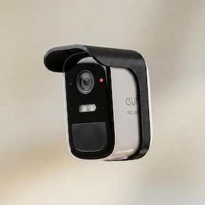 May include: A black and silver security camera with a black visor. The camera has a lens, a light, and a black panel. The word "eufy" is visible on the side. The camera is designed for home security.
