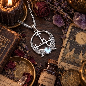 Op de afbeelding: Een zilverkleurige ronde hanger aan een ketting met een ingewikkeld zegelontwerp, een klein maansikkelaccent en een ronde iriserende cabochonsteen. De hanger hangt aan een zilverkleurige ketting en toont gedetailleerd metaalwerk en gravures.