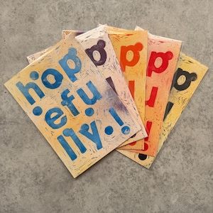 May include: A selection of handmade cards, each featuring the word "hopefully!" in various colours: blue, purple, orange, and red. The cards are fanned out, displaying the text on a beige background.