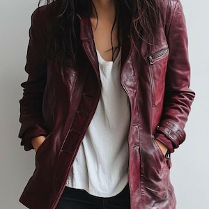 May include: A burgundy leather jacket with a button-up front, a zippered chest pocket, and side pockets. The jacket is worn over a white top and dark blue jeans. The model has long, dark hair.