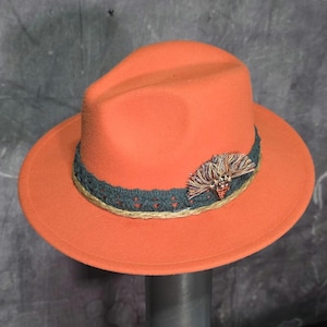 May include: A vibrant orange fedora hat with a dark teal and tan band. The hat is adorned with a decorative feather and tassel detail. The hat has a wide brim and a classic, structured shape.