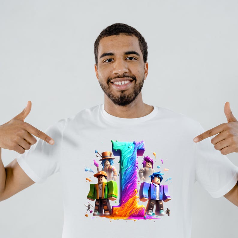 Puede incluir: Camiseta blanca con una colorida ilustraci&oacute;n del n&uacute;mero 1 y personajes de dibujos animados. El n&uacute;mero est&aacute; en tonos de azul, verde y morado, con una base naranja y amarilla. Los personajes visten diferentes atuendos.