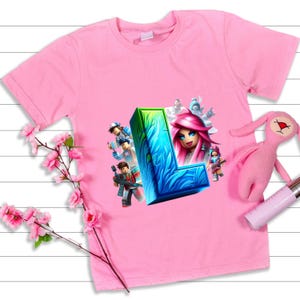 Puede incluir: Camiseta rosa de manga corta con un dise&ntilde;o gr&aacute;fico colorido. El dise&ntilde;o incluye una gran letra "L" azul y verde con ilustraciones de personajes y una mujer con pelo rosa. Un conejito de peluche rosa est&aacute; al lado de la camiseta.