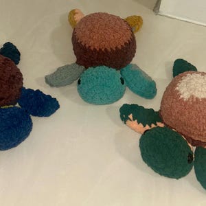 May include: Three handmade crochet turtle plushies in shades of brown, blue, teal, and green. Each turtle has a round body and flipper-like appendages. The turtles are soft and textured, perfect for a nursery or child's room.