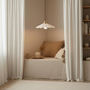 May include: A cozy reading nook with white curtains, a beige bench, and a decorative pendant light. The light fixture has a scalloped white shade and a brass-colored accent. Pillows and decorative items add to the inviting atmosphere. A rug and bookshelf are also visible.