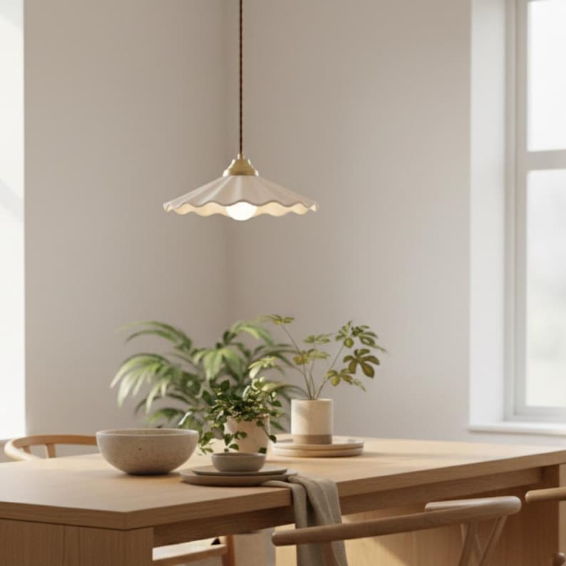 May include: A white, scalloped-edge pendant light hangs above a wooden dining table. The light fixture has a brass-colored top and a brown cord. The table is set with a bowl, plates, and greenery, creating a warm, inviting atmosphere.
