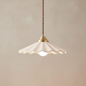 May include: A white ceramic pendant light with a scalloped edge and a brass-colored base. The light hangs from a brown, twisted cord. The light bulb is a small, round, white globe.