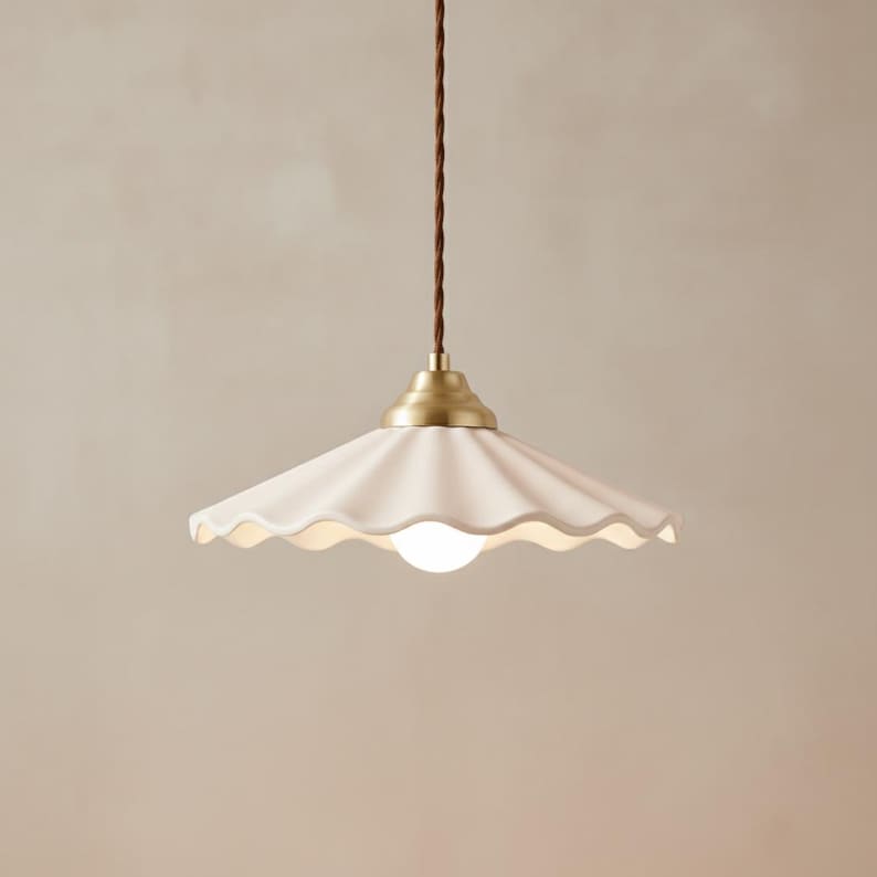May include: A white ceramic pendant light with a scalloped edge and a brass-colored base. The light hangs from a brown, twisted cord. The light bulb is a small, round, white globe.