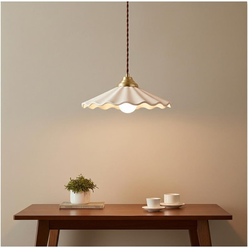 May include: A white, scalloped-edge pendant light with a brass fixture hangs from a brown cord. The light is centered above a wooden table, with a small potted plant and two teacups visible on the table. The light fixture has a retro design.