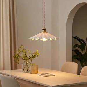 May include: A white, scalloped-edge pendant light fixture with a brass-colored base hangs above a wooden table. The light fixture is suspended by a brown cord. The room has a warm, inviting ambiance.