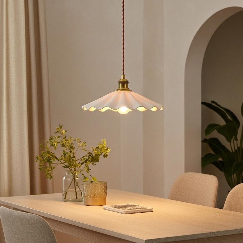 May include: A white, scalloped-edge pendant light fixture with a brass-colored base hangs above a wooden table. The light fixture is suspended by a brown cord. The room has a warm, inviting ambiance.