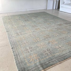 May include: A rectangular, light blue rug with a floral pattern. The rug has a border and is made of a soft material. The floral pattern is in shades of beige and gray.