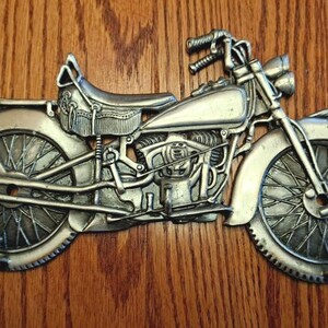 May include: A silver-colored motorcycle wall hook with intricate detailing. The vintage-style motorcycle features a seat, fuel tank, and spoked wheels. The hook is mounted on a wooden surface.