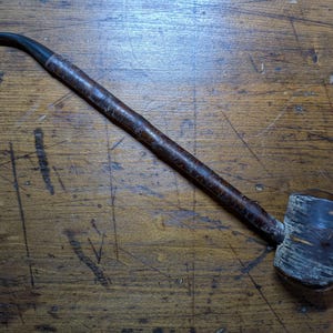 May include: A vintage tobacco pipe with a long, dark brown stem and a rounded bowl. The bowl has a weathered, textured appearance. The pipe rests on a wooden surface with visible grain and dark markings.