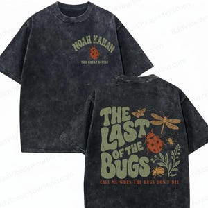Op de afbeelding: Een antracietgrijs acid-wash t-shirt met een kleine afbeelding op de voorkant van een lieveheersbeestje en de tekst Noah Kahan The Great Divide. De achterkant toont grote groene tekst The Last of the Bugs met illustraties van insecten.