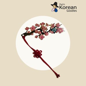 May include: A Korean-style ornament with a branch of pink flowers and a blue butterfly. A red knotted cord hangs below. The text "Park's Korean Goodies" is in the upper right corner.