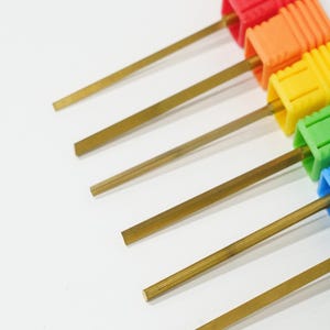 May include: A set of six metal tools with long, thin, gold-colored shafts and square-shaped handles in a rainbow gradient of red, orange, yellow, green, blue, and purple. Each tool features a flat, straight metal tip extending from the handle.