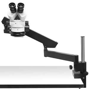 May include: A stereo microscope with a white and black body, featuring dual eyepieces and adjustable arm. The microscope is mounted on a black articulated arm, attached to a white surface with a clamp.