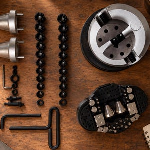 May include: A jewelry engraving vise set featuring a large circular silver-toned vise, two silver-toned conical attachments, two long rows of small black circular components, a black T-handle hex wrench, and a black base plate with various metal pins.