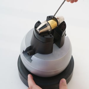 May include: A silver and gold metal object is held in a black and gray vise. A hand holds a metal tool, possibly a file, near the object. The vise sits on a black base.