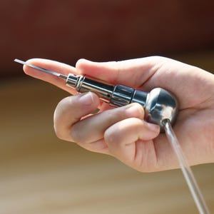 May include: A silver-colored metal tool with a thin, flat, pointed blade extending from a cylindrical handle. The handle features a rounded, bulbous end and a clear, flexible plastic tube extending from the base of the metallic component.