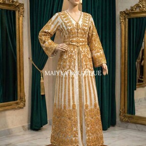 May include: A white and gold embroidered gown on a mannequin. The long-sleeved dress features intricate gold patterns, a V-neck, and a matching belt. The dress is displayed on a wooden platform in front of a green curtain and gold-framed mirrors.