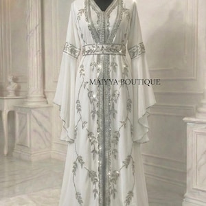 Peut inclure: Longue robe blanche avec des manches évasées et une broderie argentée détaillée. La robe présente un décolleté en V, une taille cintrée et des motifs de feuilles. Le texte "MAIYYA BOUTIQUE" est visible.