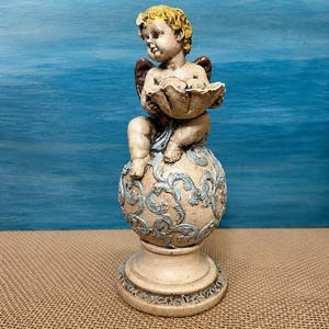 Könnte beinhalten: Eine dekorative Figur eines Cherubs mit goldenem Haar und kleinen Flügeln, der eine muschelförmige Schale hält. Der Cherub sitzt auf einer kugelförmigen Basis mit hellblauen Rankenmustern, die auf einem runden Sockel ruht.