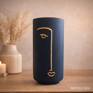 Può includere: Un vaso cilindrico blu navy scuro caratterizzato da un disegno a linea dorata continua che raffigura il profilo di un volto umano. Il design include un occhio semplice, una linea del naso dritta e una piccola bocca. Il testo ATÖYLE IZGI è visibile in basso a destra.