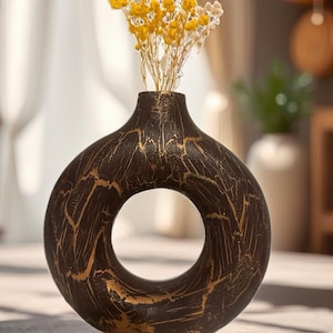 Könnte beinhalten: Dekorative, dunkelbraune Vase mit kreisförmiger Öffnung und goldenen Akzenten. Sie enthält einen Strauß getrockneter gelber und weißer Blumen. Die Vase steht auf einer Holzoberfläche.