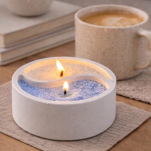 Puede incluir: Una vela redonda blanca con un diseño de yin-yang, llena de arena amarilla y azul, y dos mechas encendidas. Una taza beige de café está cerca sobre un posavasos tejido. La vela está sobre una mesa de madera.