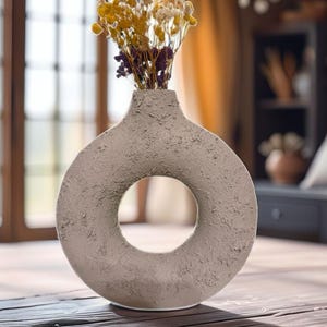 以下が含まれることがあります： ベージュ色のリング型花瓶。表面はざらざらとした質感で、黄色と紫色のドライフラワーが入っています。花瓶の中央には円形の穴があり、木製のテーブルの上に置かれています。インテリア用の装飾品です。