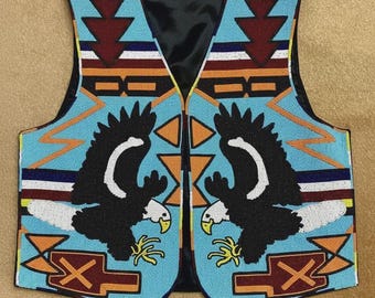 Puede incluir: Un chaleco con cuentas con forro negro y fondo azul claro. El chaleco presenta dos águilas negras con detalles blancos enfrentadas. Patrones geométricos en rojo, naranja y marrón adornan el diseño del chaleco. Es una prenda tradicional nativa americana.
