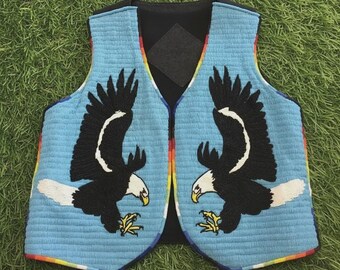 Puede incluir: Un chaleco azul claro con un diseño de cuentas que presenta dos águilas con alas negras, cabezas blancas y picos amarillos. Los bordes están adornados con un patrón de arcoíris. El chaleco se muestra sobre una superficie verde.