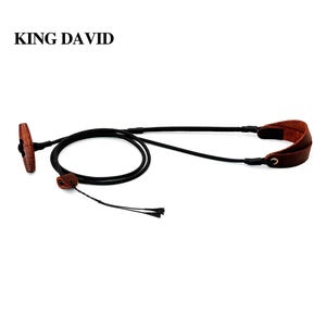 Puede incluir: Una correa de cordón negro con un pasador de madera marrón y detalles de cuero marrón. El texto KING DAVID está impreso en negro en la esquina superior izquierda. La correa presenta un diseño de bucle con un ajustador de cuero y dos extremos.
